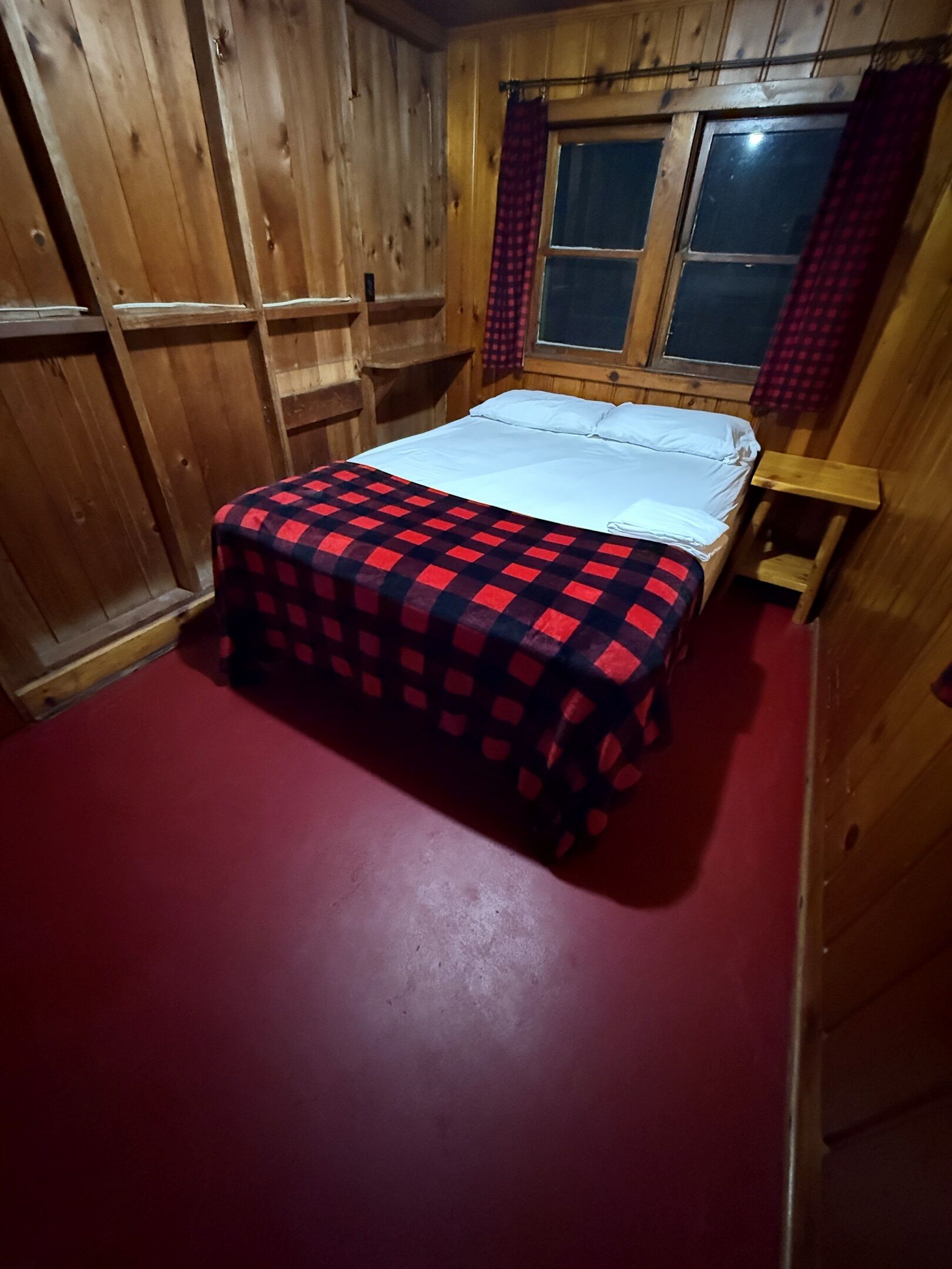 Una habitación de madera con una cama, un ventanal y un suelo rojo.