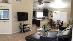 Uma sala de estar com sofá de couro preto, televisão, pequena mesa de centro e área de jantar com cadeiras e mesa.