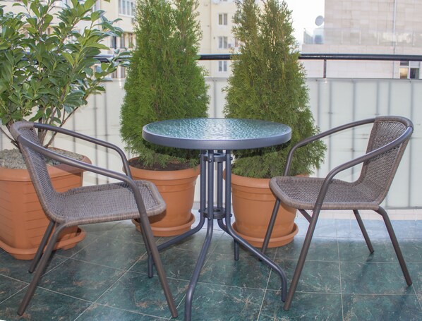 A small outdoor seating area with two wicker chairs and a glass-top table, surrounded by potted plants.