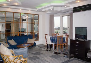 A modern living room with a blue sofa, wooden chairs, a dining table, and a TV.