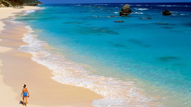 A beach with clear turquoise waters and a person walking along the shore. - Cottage Priv. Pool & Tennis Beach 5 min Central (Paget)