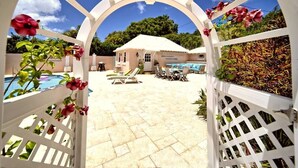 A pool area with a white pergola, lounge chairs, and a small building in the background. - Cottage Priv. Pool & Tennis Beach 5 min Central (Paget)