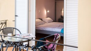 A balcony with a table and chairs, a bed with white bedding, and a wall-mounted light fixture. - Lourdas bay great view - Studio Maria (Kefalonia)