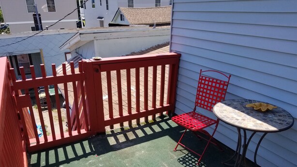 A small outdoor patio with a red metal fence, a blue patterned rug, a round table, and two red chairs.