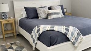 Quarto com cama branca, roupa de cama azul escura, tapete estampado e janela com três plantas em vasos.
