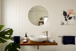 Ein Badezimmer mit Waschtisch aus Holz, einem weißen Waschbecken, einem runden Spiegel und einem Handtuchhalter an der Wand.