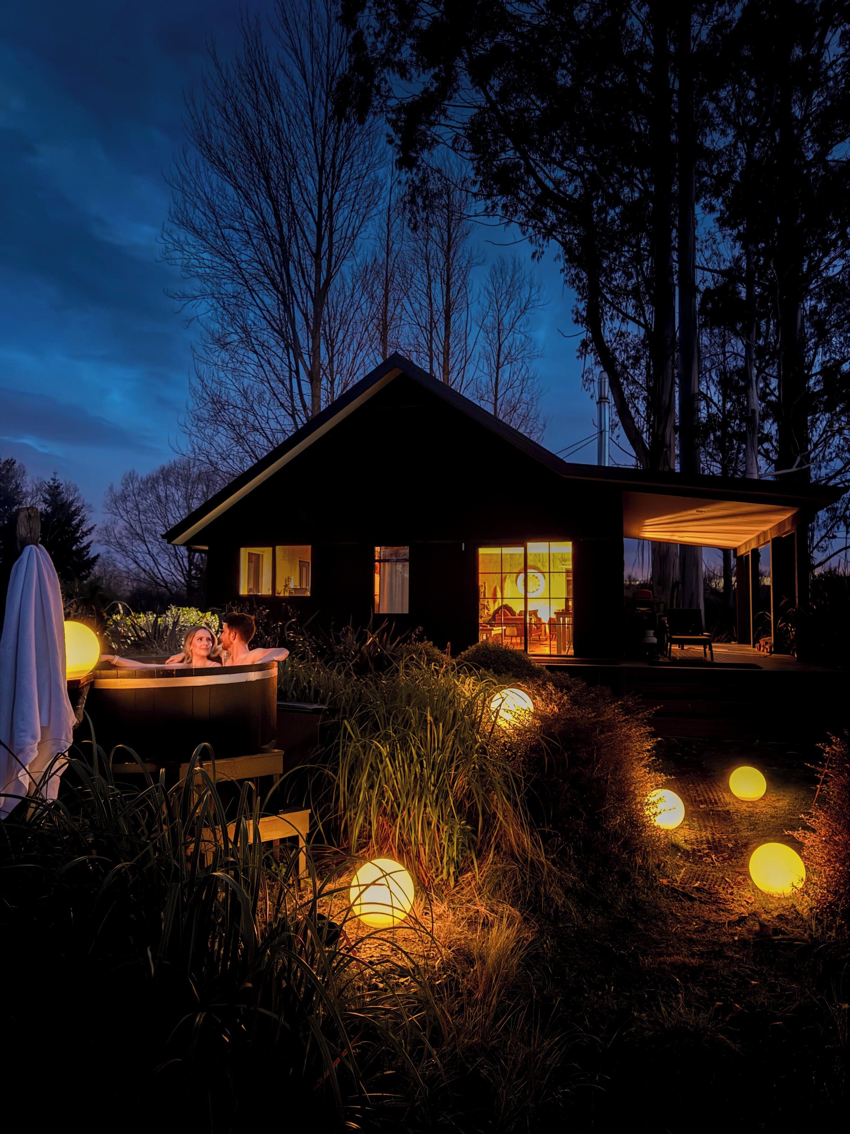 Ein modernes Haus mit Whirlpool, umgeben von hohen Bäumen und beleuchtet mit kugelförmigen Lichtern.