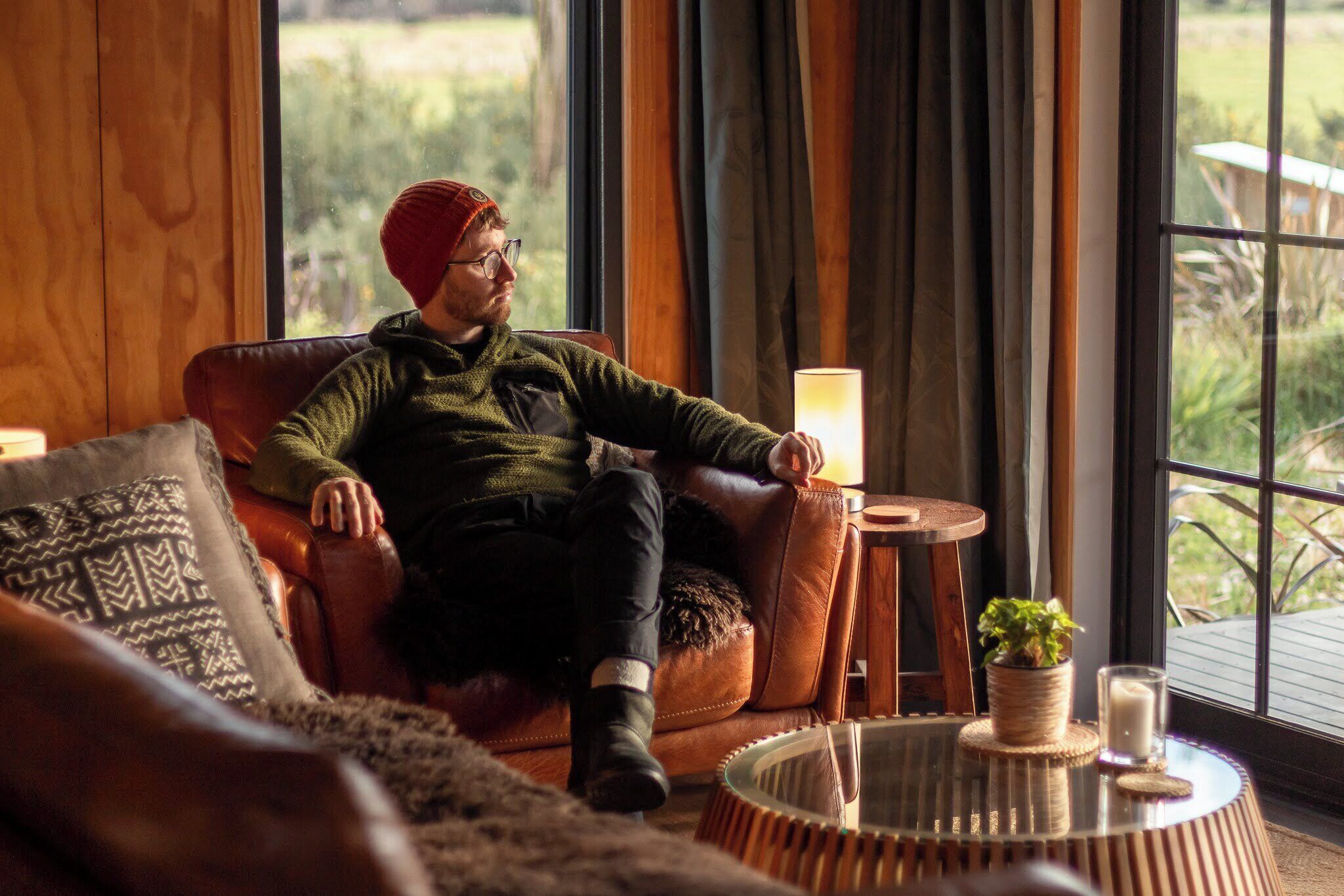 A person seated on a leather sofa in a room with a view of greenery outside. There is a round wooden table with a lamp and a small plant.