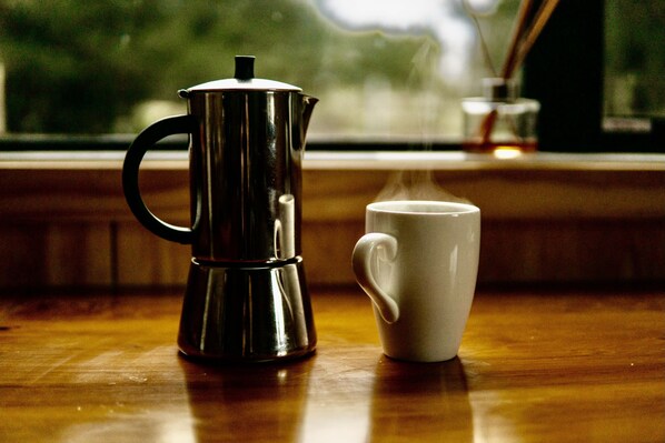 Ein Edelstahl-Kaffeekocher und eine weiße Tasse auf einem Holztisch in der Nähe eines Fensters.