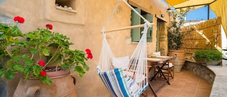 A hammock hanging from a rope, a potted plant with red flowers, and a table with chairs outside a building with a yellow awning.