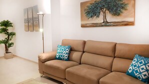 Un salon avec un canapé beige, deux coussins à motifs, une lampe sur pied, une plante en pot et un tableau représentant un arbre.