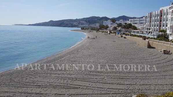 Ein Strand mit einem gepflasterten Weg, einem Hotelgebäude und einem Berg im Hintergrund.