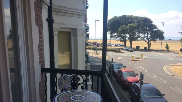 Un balcón con mesa y sillas de cerámica, con vista a una calle con coches estacionados y al mar.