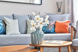 A living room with a grey sofa, a blue vase with white flowers, and a round wooden table with a magazine.