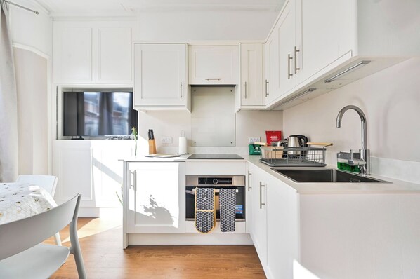 Cocina compacta con muebles blancos, horno empotrado, pileta de acero inoxidable y ventana que proporciona luz natural.