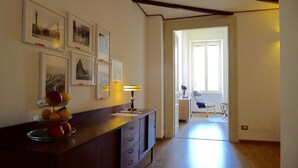 Ein Zimmer mit einem Holztisch, einer Lampe und einer Obstschale. An der Wand hängen Bilder in Rahmen, und eine Tür führt in einen anderen Raum.