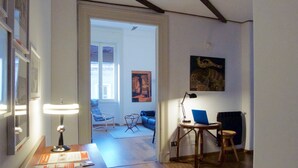 Ein Hotelzimmer mit Holzboden, einem Schreibtisch mit Lampe, einem Laptop, einem Sessel und einem Sofa. Eine Tür führt in einen anderen Raum.
