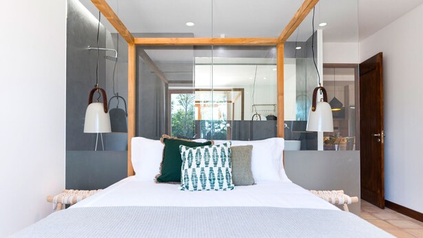 Een moderne slaapkamer met een groot bed, een houten balkenplafond en uitzicht op de badkamer door een glazen wand.