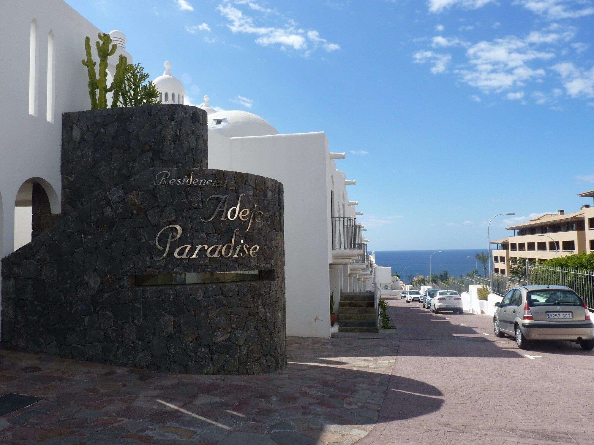 A stone entrance with "Residencial Adeje Paradise" sign, a white building with arches, and a view of the sea and parked cars.