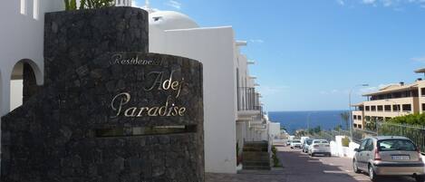 A stone entrance with "Residencial Adeje Paradise" sign, a white building with arches, and a view of the sea and parked cars.