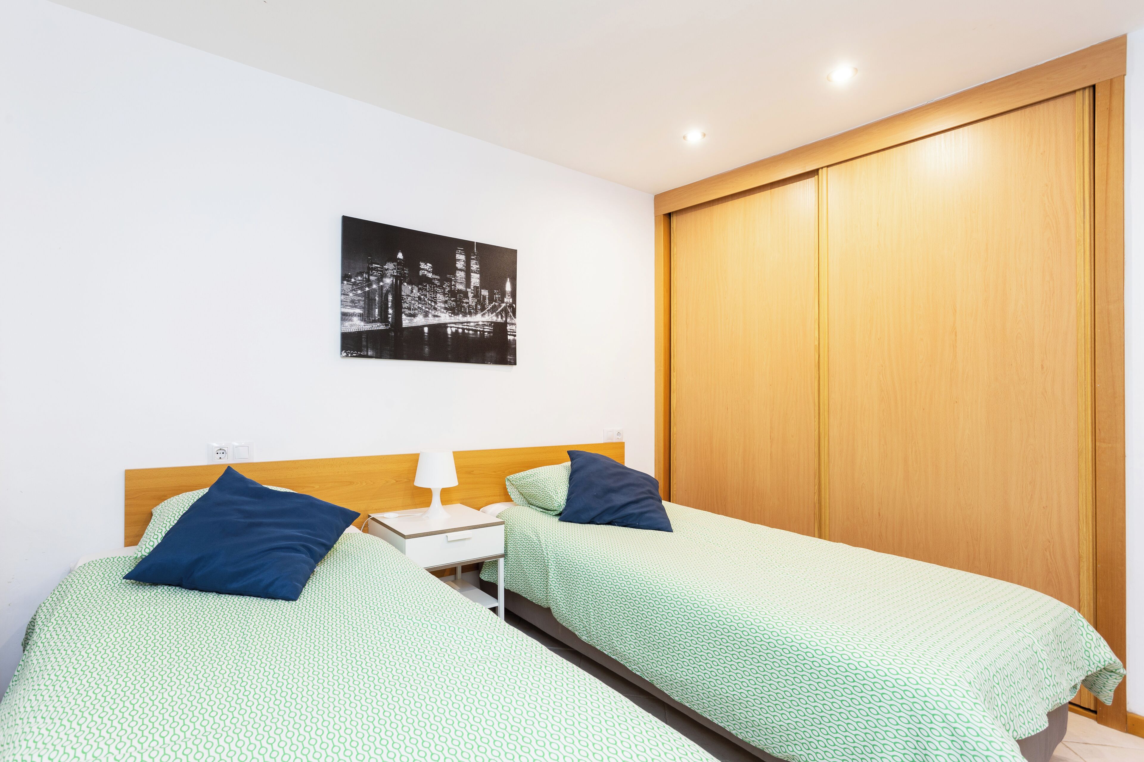 Een kamer met twee bedden, een houten garderobe, een nachtkastje, een lamp en een stadsgezicht aan de muur.
