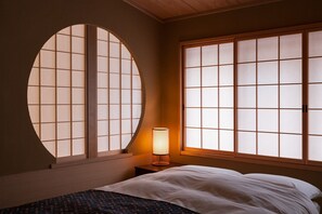 Traditional Japanese-style bedroom with a bed against a wall, featuring a large window and a bedside table with a lamp.
