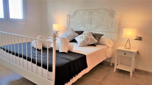 Una cama bien hecha con almohadas y una manta, una mesita de noche con lámpara, y un cabecero blanco.