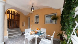 Un patio con una mesa lista para el desayuno, sillas de mimbre blanco, piso de piedra, un portón de madera y una planta en maceta.