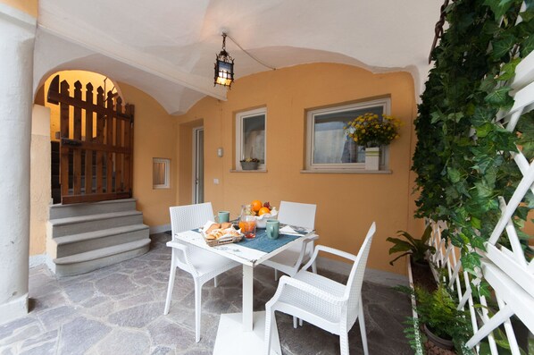A covered outdoor seating area with a table set for two, surrounded by white wicker chairs, a stone floor, and a view of a building entrance with a wooden gate.