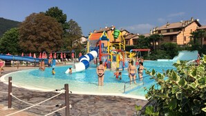 Zona de piscina con tobogán y estructuras de juegos, rodeada de sillones y sombrillas.