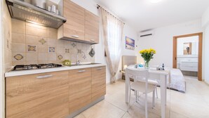 Een kleine keuken met houten kastjes, een gasfornuis, en een eethoek met een tafel en stoelen.