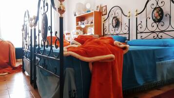Un dormitorio con estructura de cama metálica, colcha azul, manta roja y mesita de noche de madera con una lámpara.