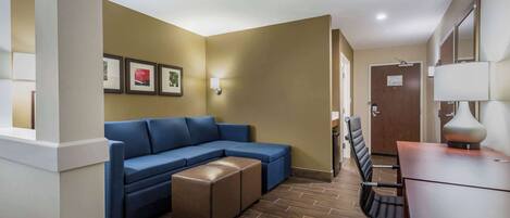 A hotel room with a blue sectional sofa, a wooden desk, and a chair. There are framed pictures on the wall and a door in the background.
