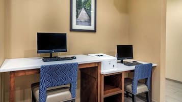 Um espaço de trabalho com duas mesas, cada uma com um computador e teclado. Há duas cadeiras com capas estampadas. Uma moldura com uma imagem está pendurada na parede acima das mesas.