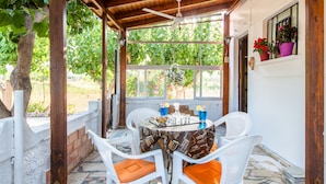 A covered patio with a table set for two, white chairs with orange cushions, and a view of greenery through the windows. - Villa Aadonis in the sea (Chania kolibari)