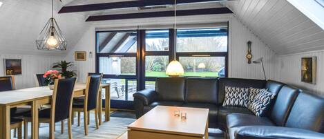 A cozy living room with a wooden table, chairs, a sofa, and a coffee table. There is a large window with a view of the outdoors.