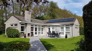 A two-story house with a brick exterior, large windows, a covered patio with outdoor furniture, and a solar panel on the roof. - Gravenhof 12 bungalow on holiday park in forest and dune area (Burgh-Haamstede)