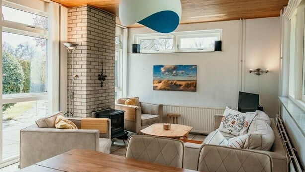 A cozy living room with a fireplace, comfortable seating, a dining table, and a large window offering a view of the outdoors. - Gravenhof 12 bungalow on holiday park in forest and dune area (Burgh-Haamstede)