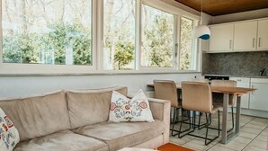 A modern living room with a beige sofa, patterned cushions, and a wooden coffee table. There are large windows offering a view of trees and a kitchen area with white cabinets and a hanging light fixture. - Gravenhof 12 bungalow on holiday park in forest and dune area (Burgh-Haamstede)