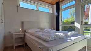 A bedroom with a bed, a nightstand, a window, and a view of trees. - Gravenhof 12 bungalow on holiday park in forest and dune area (Burgh-Haamstede)