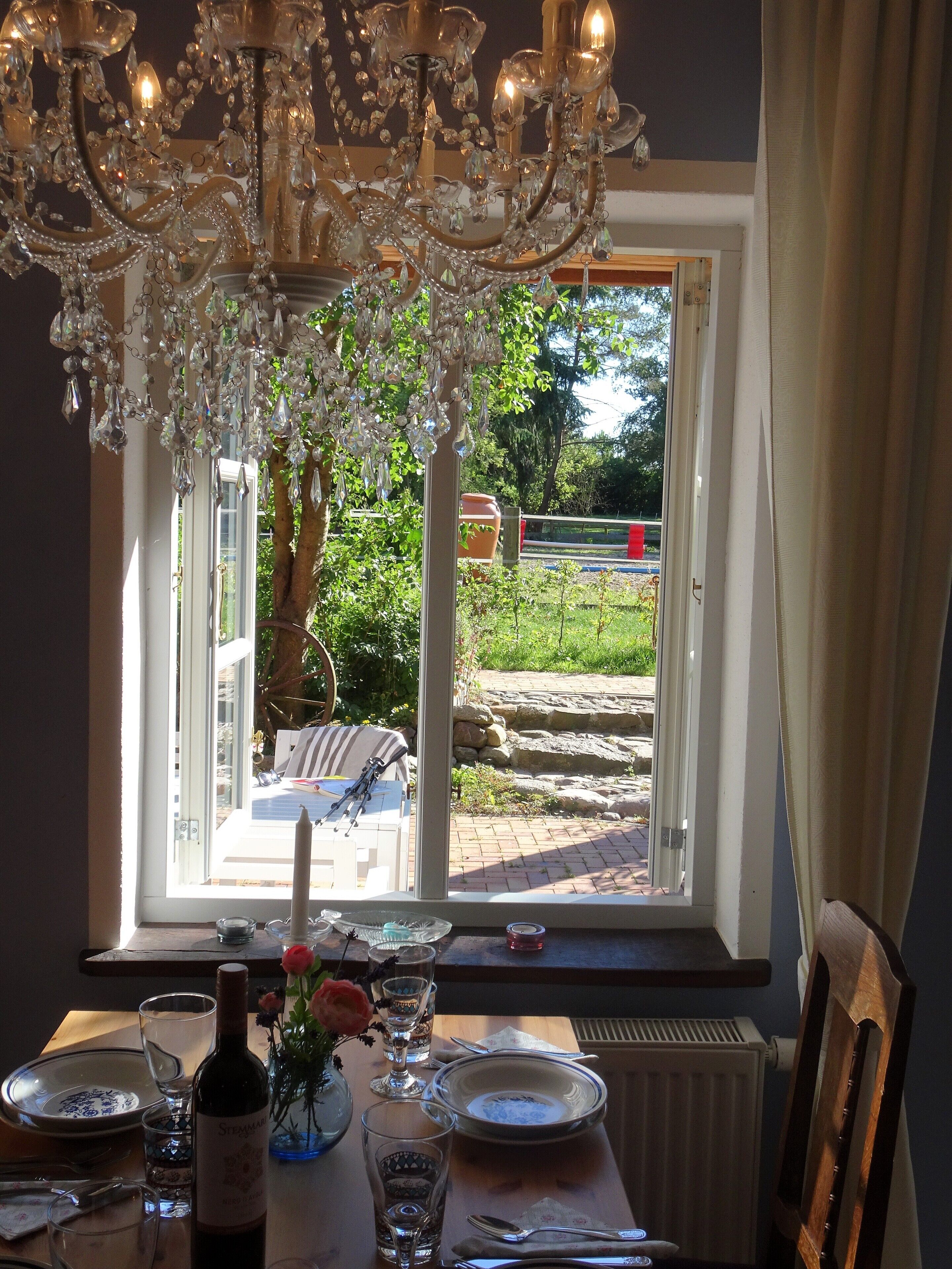 A dining table set with plates, glasses, and a bottle, with a chandelier above and a view of a garden outside.