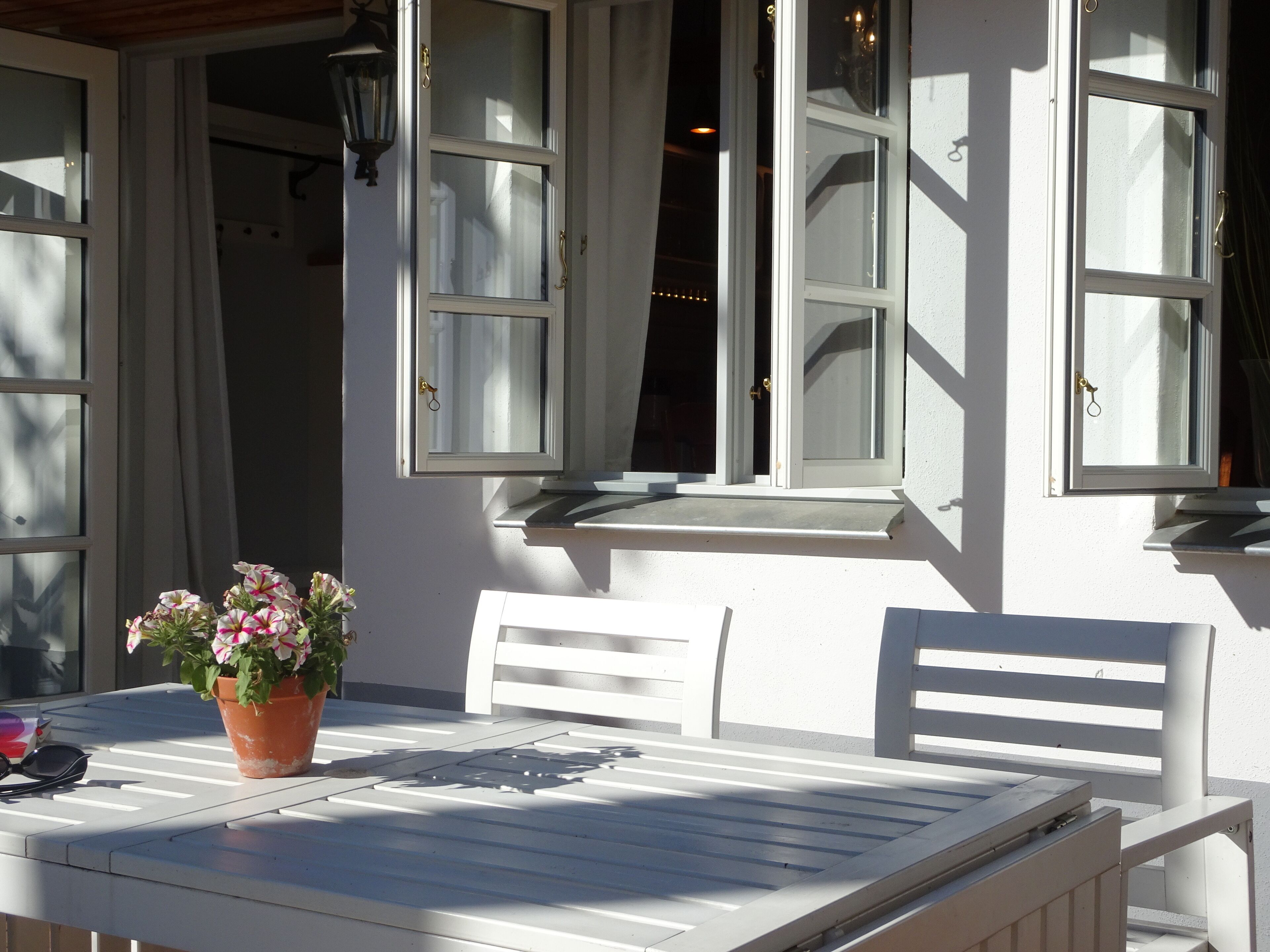 A white table with a potted plant and sunglasses on it, a white chair, and a window with open shutters.