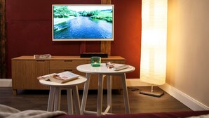 Ein gemütliches Zimmer mit Fernseher an der Wand, einem kleinen Tisch mit Buch, Kerze und Fernbedienung.