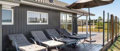 A deck with four lounge chairs and a large umbrella, attached to a building with a tiled roof and windows.
