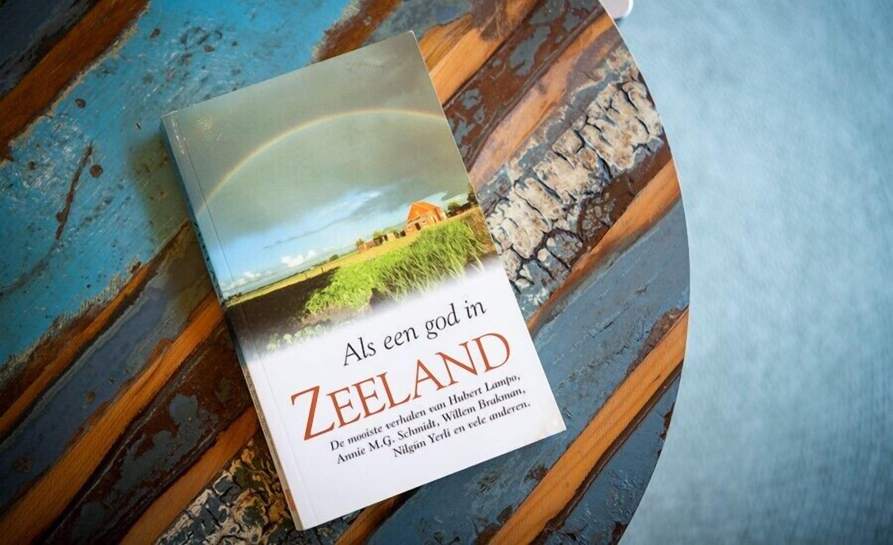Un libro titulado "Als een god in Zeeland" con una imagen de paisaje en la portada, sobre una superficie de madera con un diseño azul y marrón.