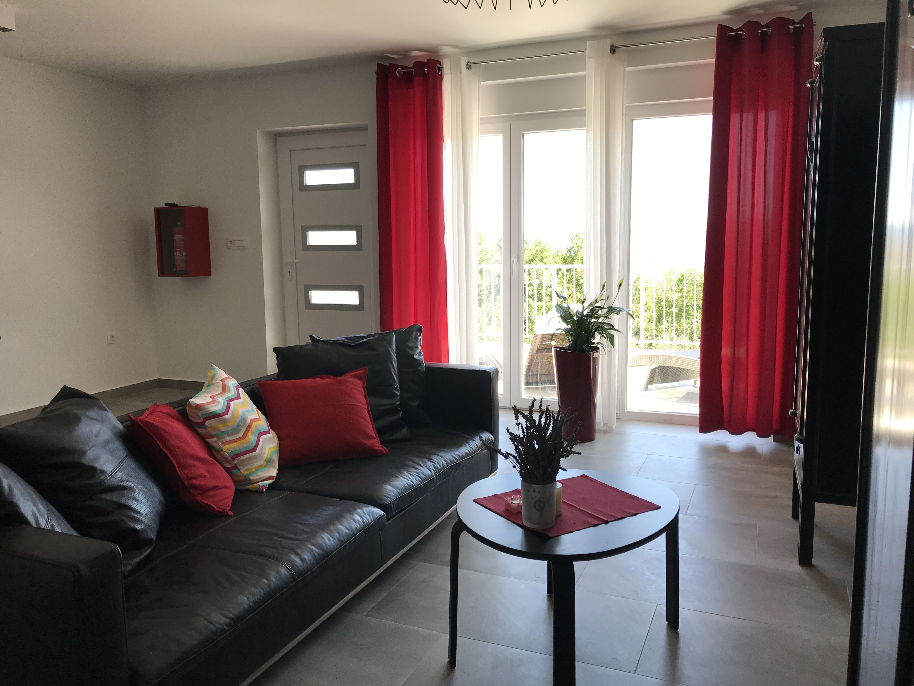 Ein Wohnzimmer mit einer schwarzen Ledersofa, roten und bunten Kissen, einem kleinen runden Tisch mit einer Vase und einer Pflanze, und Blick auf einen Balkon mit roten Vorhängen.