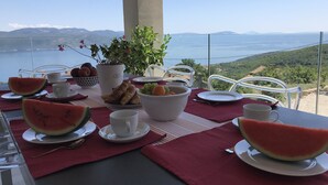 Ett bord dukat med en vattenmelon, croissanter, vindruvor och en skål med frukt, med utsikt över ett vackert landskap med en sjö eller ett annat vattendrag och kullar.