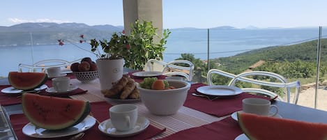 Ett bord dukat med en vattenmelon, croissanter, vindruvor och en skål med frukt, med utsikt över ett vackert landskap med en sjö eller ett annat vattendrag och kullar.