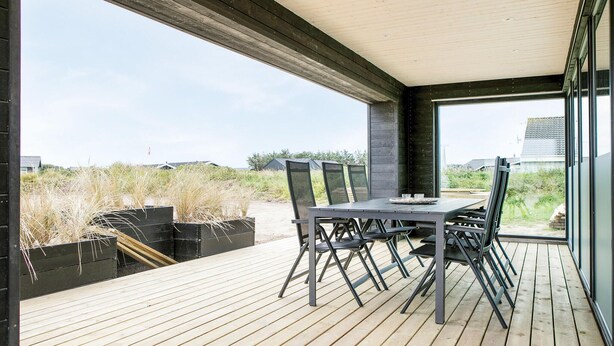 An outdoor dining area with a table and chairs, a wooden deck, and a view of the beach and buildings in the distance.