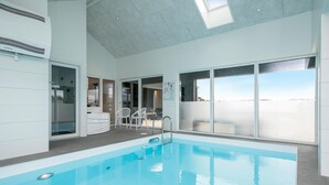 A modern indoor swimming pool with clear glass walls, a ceiling with recessed lighting, and a view of the water outside.